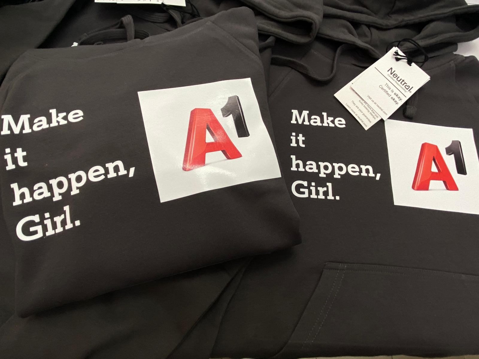 Folded black hooded sweatshirts with a white printed slogan and a printed square logo, still carrying their garment tags.