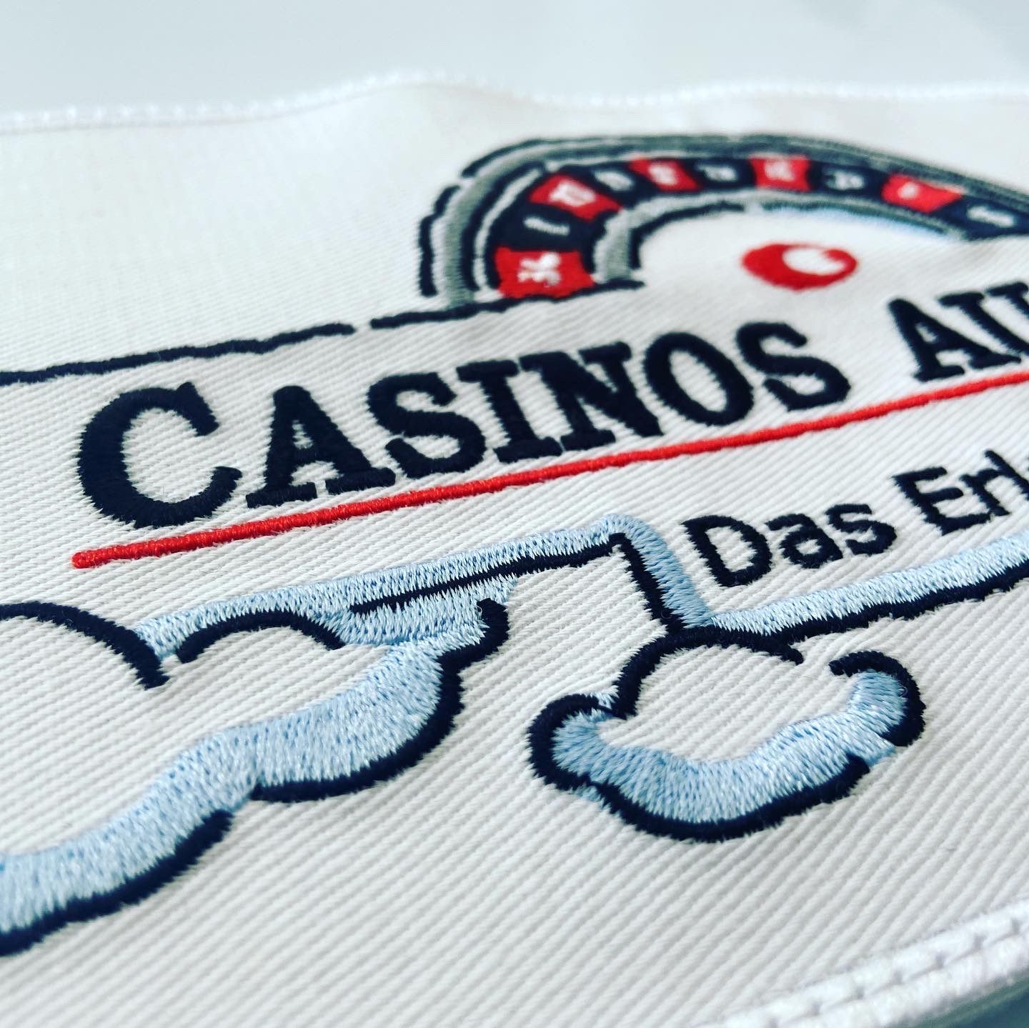Macro view of a white embroidered patch with dark blue lettering, a red line and light blue outlines, showing individual satin stitches.