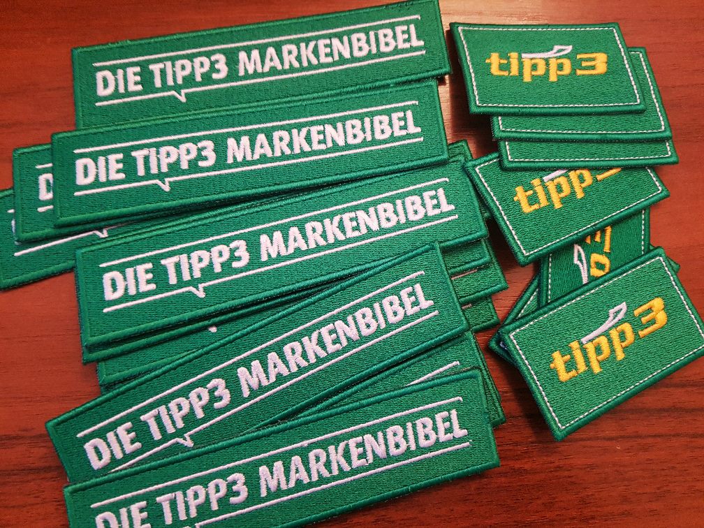 Stacks of green fully embroidered patches in two sizes, with white and yellow lettering, spread on a wooden table.
