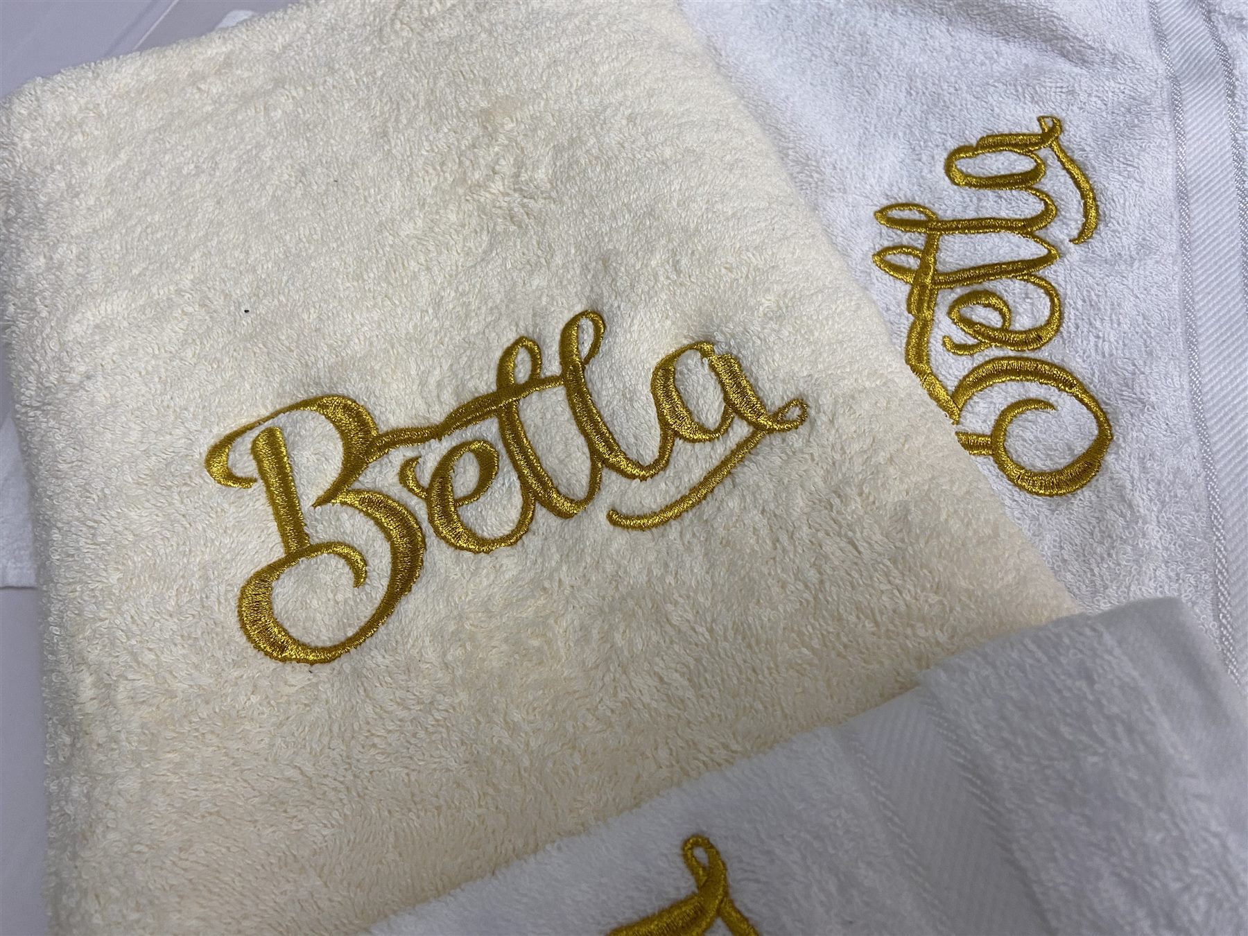 Folded cream and white terry towels, each embroidered with a different first name in gold script lettering.