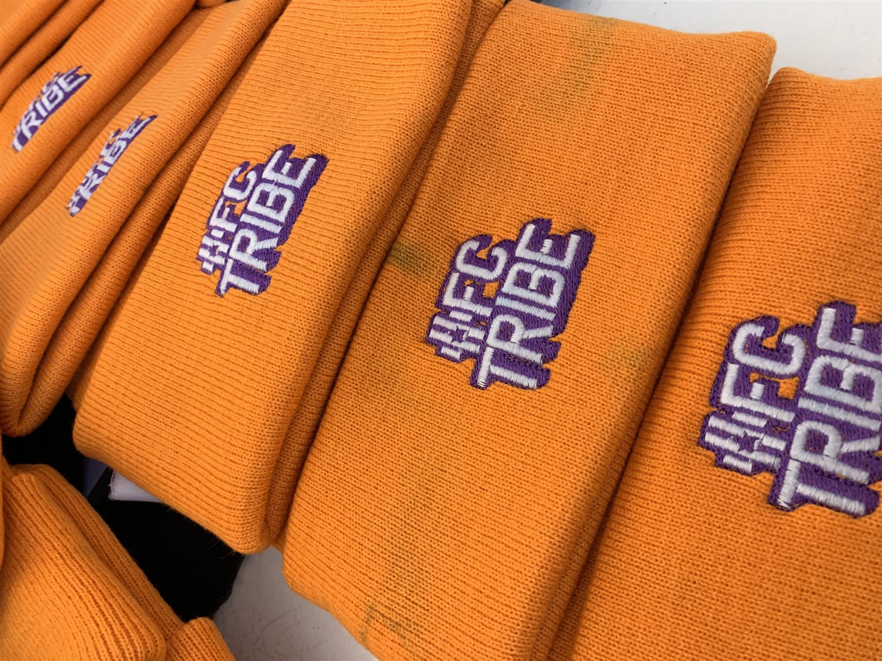 Row of folded orange knitted beanies, each embroidered with the same purple and white FC Tribe logo on the turn-up.