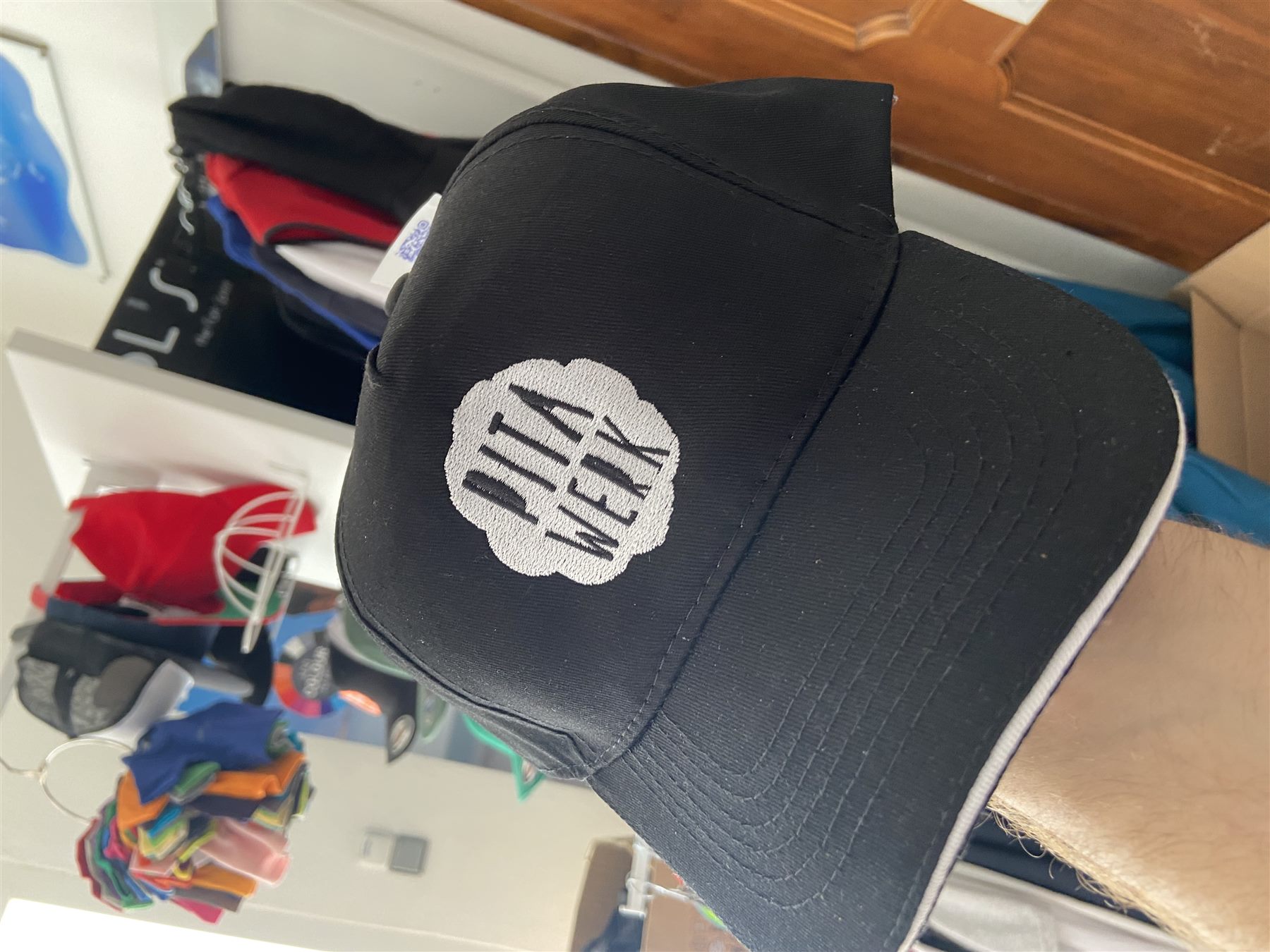 Black baseball cap with a round white embroidered logo badge on the front panel, photographed in the workshop.