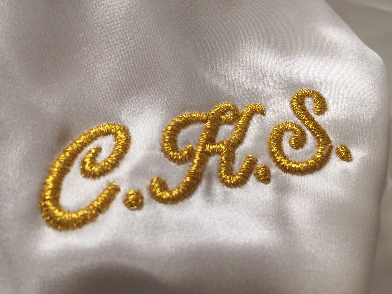 Gold metallic script monogram with three initials embroidered on white satin fabric.
