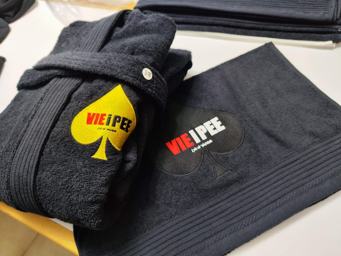 Dark bathrobe and folded dark towel, each embroidered with a spade-shaped VIE i PEE logo in yellow and in tonal thread.