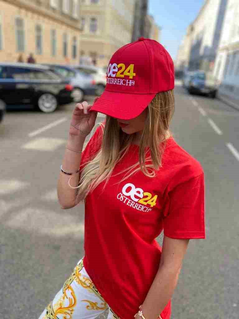 Woman on a Vienna street wearing a red cap and a red t-shirt, both carrying the same embroidered oe24 Österreich logo.
