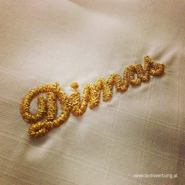 Close-up of a first name embroidered in gold metallic thread in a flowing script on cream linen.